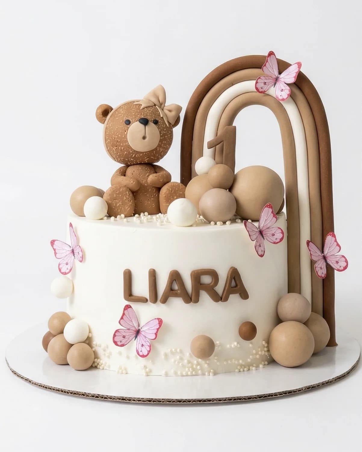 Neutral boho first birthday cake with a teddy bear, rainbow arch and butterflies, a custom cake by Cakes By Vjollca in Pristina, Kosovo
