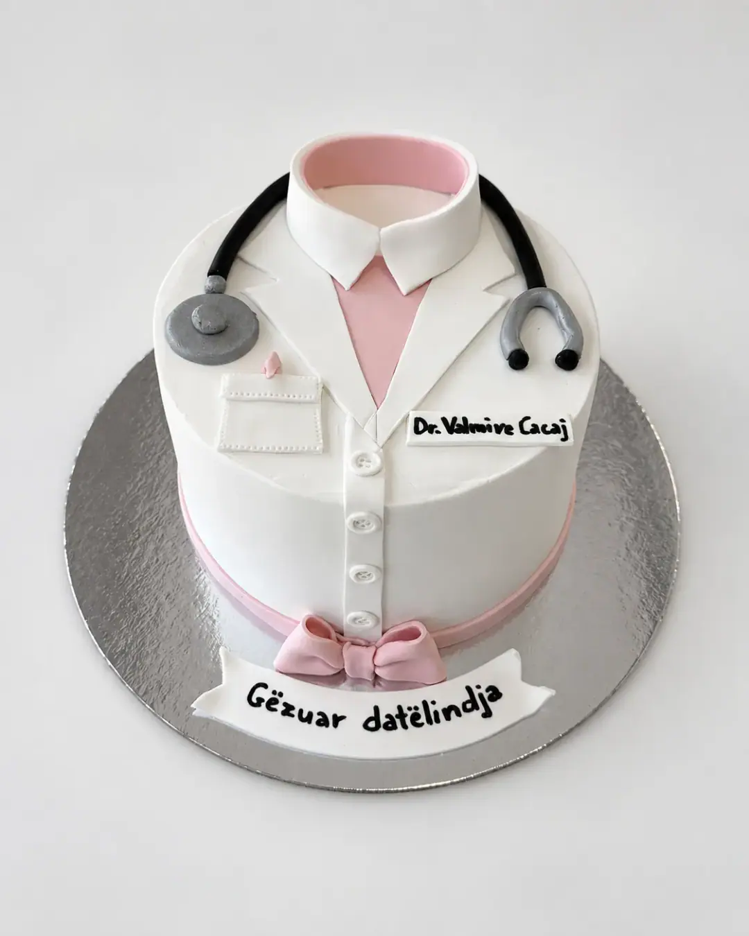 White birthday cake dressed as a doctor's coat with a pink shirt collar, fondant stethoscope, name badge, buttons and pink bow, and a 'Gëzuar ditëlindja' banner, a custom cake by Cakes By Vjollca in Pristina, Kosovo