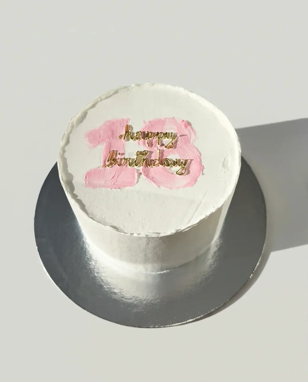 White buttercream birthday cake with a brushed pink 18 on top and 'happy birthday' in raised gold script, a custom cake by Cakes By Vjollca in Pristina, Kosovo