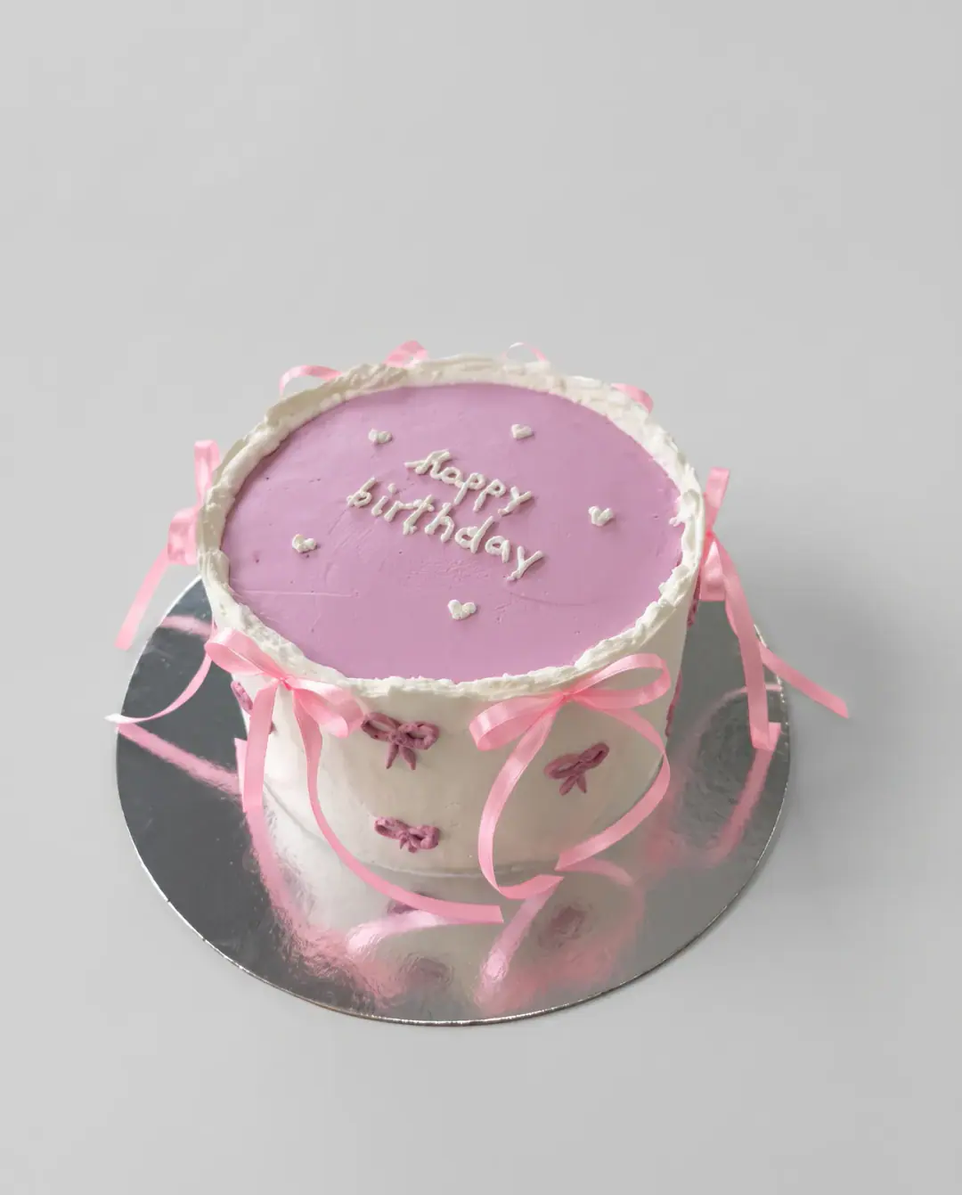 White vintage birthday cake with a lilac top, 'happy birthday' in white script, pink satin ribbons tied around the sides and small piped bows, a custom cake by Cakes By Vjollca in Pristina, Kosovo
