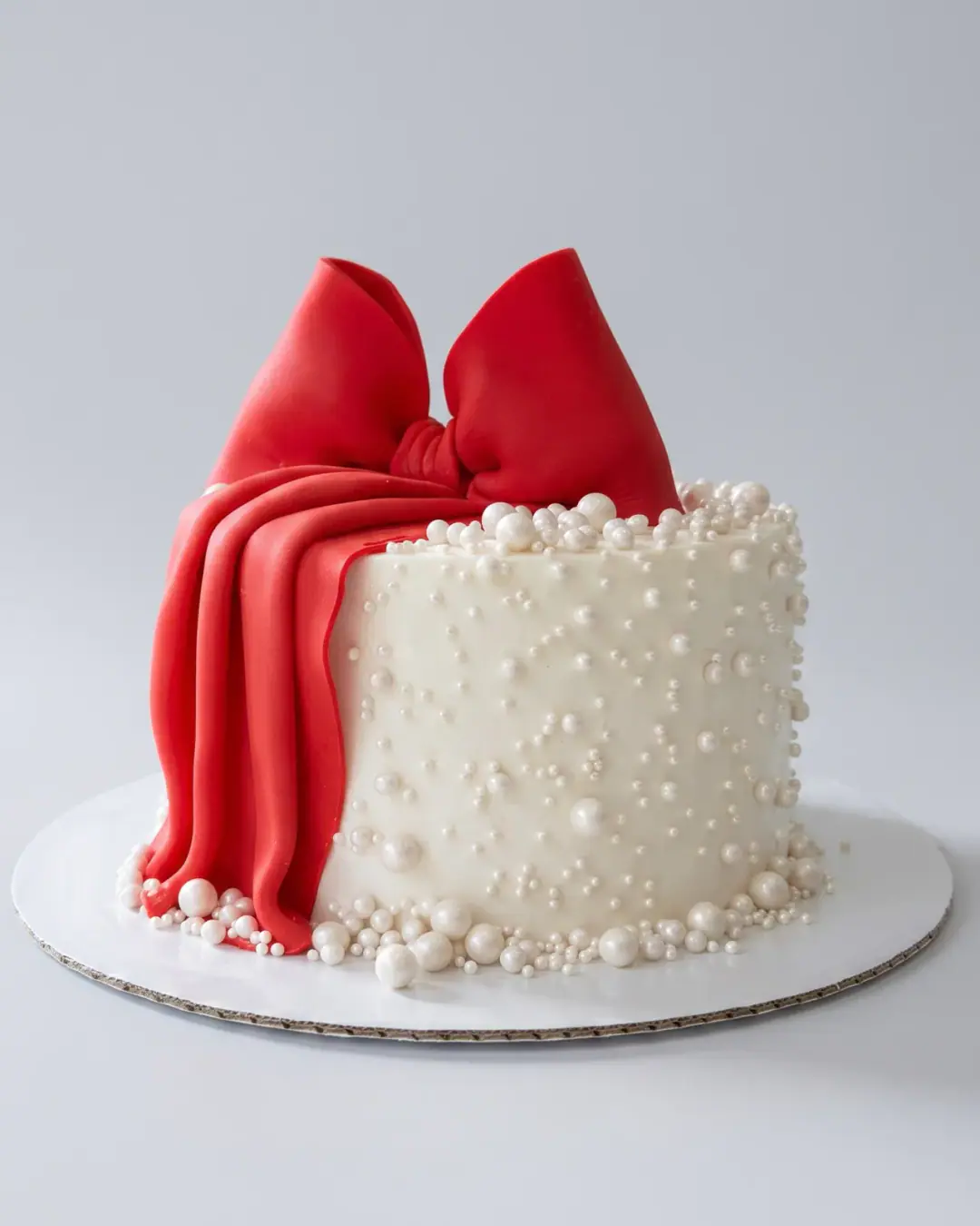 White birthday cake covered in sugar pearls with a large red fondant bow draped down one side, a custom cake by Cakes By Vjollca in Pristina, Kosovo
