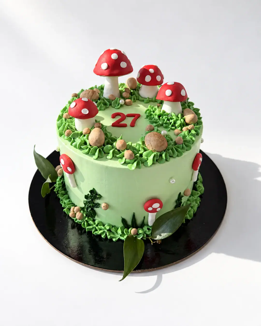 Pale green birthday cake with red and white toadstools, piped grass, small brown pebbles and a red 27 on top, a custom cake by Cakes By Vjollca in Pristina, Kosovo