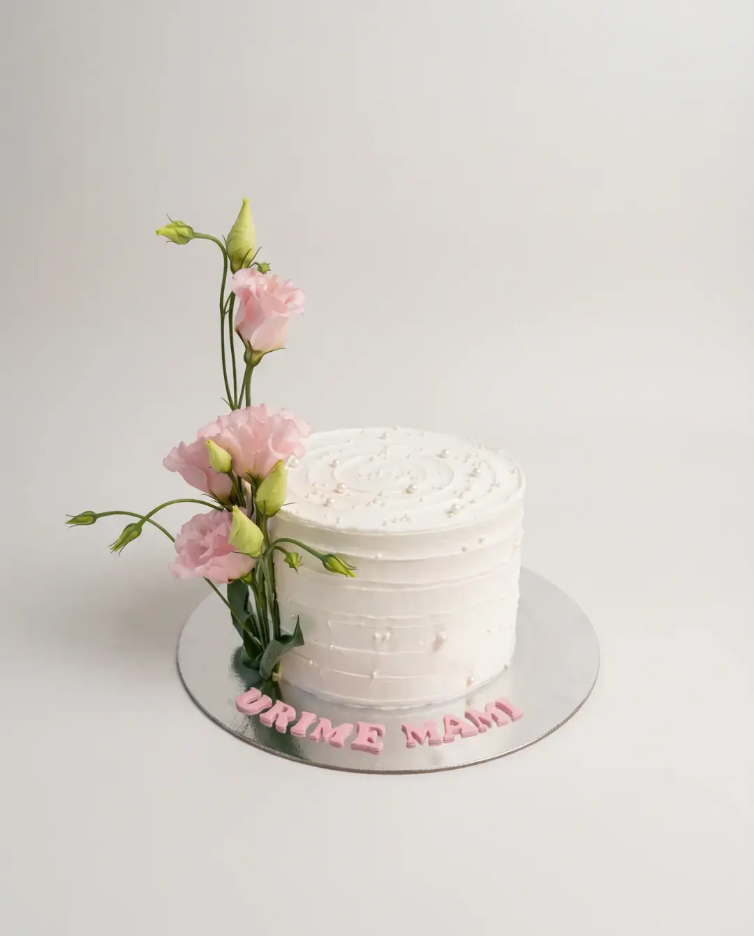 White textured buttercream cake with a tall spray of fresh pink lisianthus at one side and 'Urime Mami' in pink letters on the board, a custom cake by Cakes By Vjollca in Pristina, Kosovo