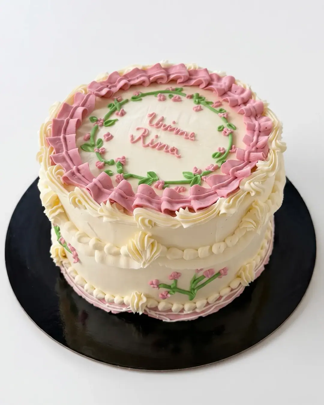 Cream vintage-style birthday cake with dusty pink ruffled piping, a piped floral wreath and hand-written lettering, a custom cake by Cakes By Vjollca in Pristina, Kosovo
