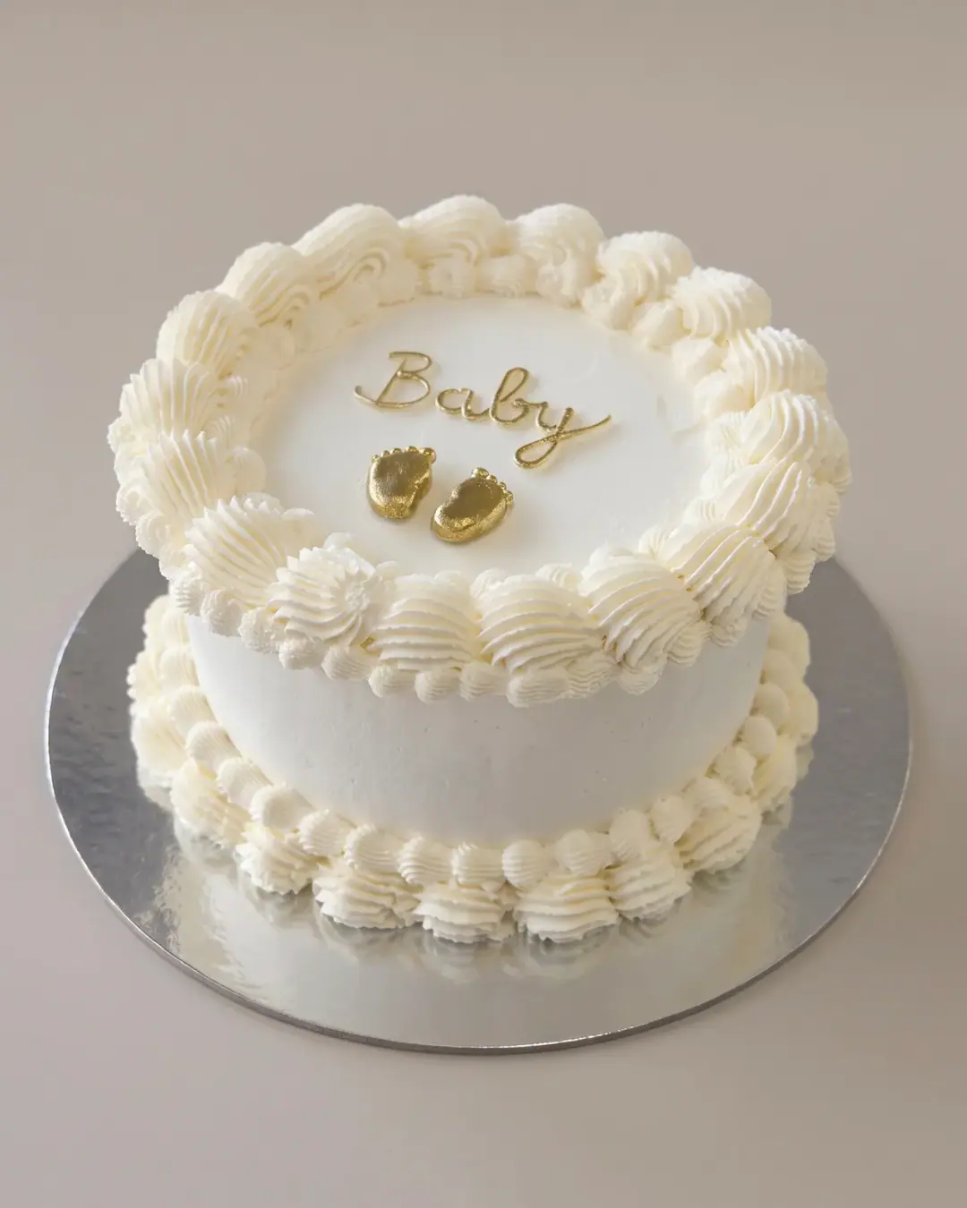 White vintage-style celebration cake with 'Baby' in gold script and two gold baby feet, a custom cake by Cakes By Vjollca in Pristina, Kosovo