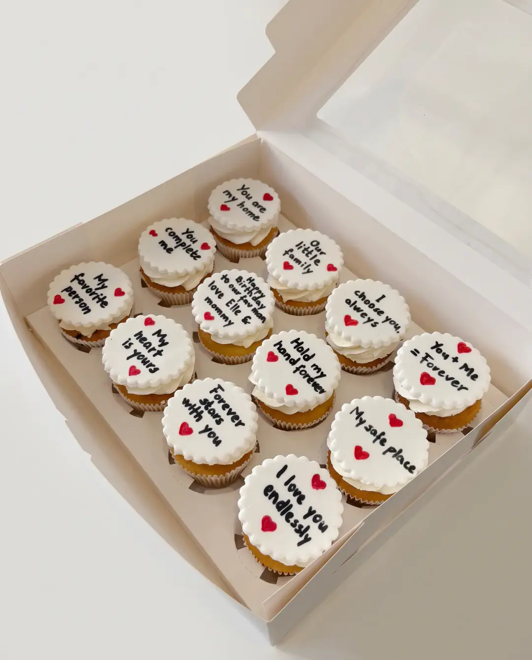 Kuti me dymbëdhjetë cupcake me disqe të bardha fondani me mesazhe dashurie të shkruara me dorë dhe zemra të kuqe, cupcake me porosi nga Cakes By Vjollca në Prishtinë