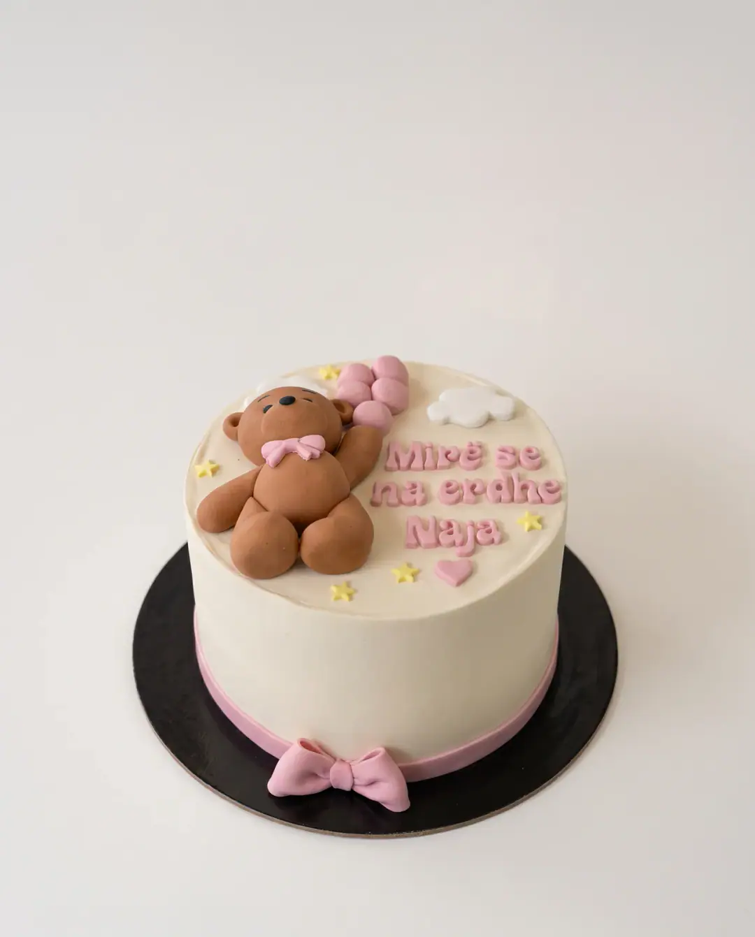 Torte krem për mirëseardhjen e një bebeje, me arush prej fondani që mban tullumbace rozë, urimin 'Mirë se na erdhe' dhe emrin e bebes në rozë, dhe shirit e fjongo rozë në bazë, torte me porosi nga Cakes By Vjollca në Prishtinë