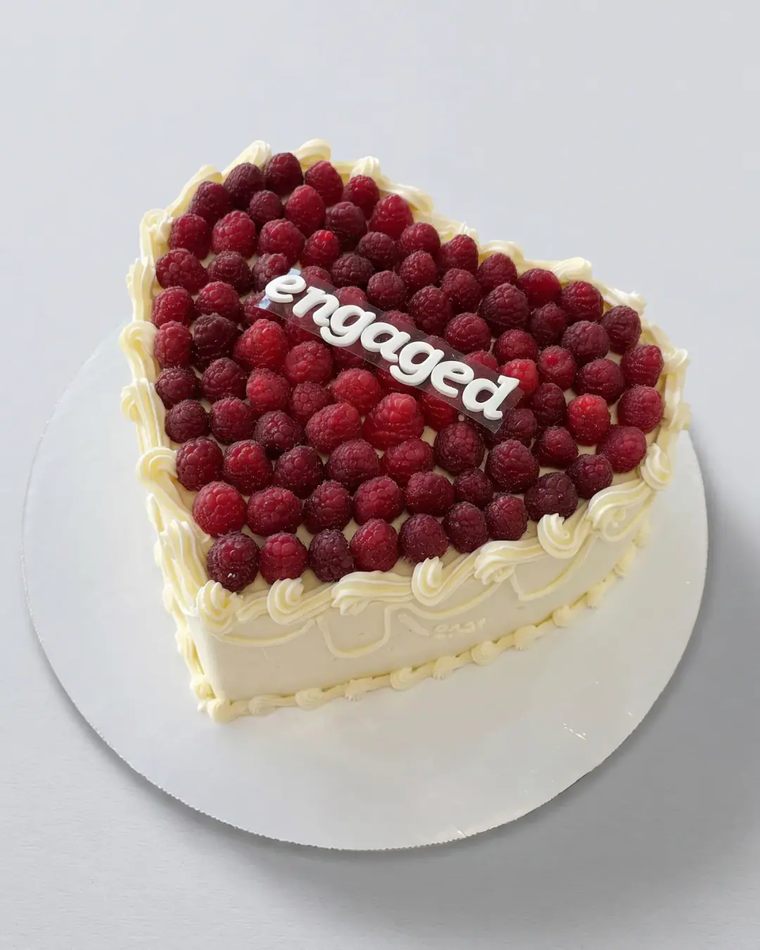 Heart-shaped engagement cake covered in fresh raspberries with cream piping and an 'engaged' plaque, a custom cake by Cakes By Vjollca in Pristina, Kosovo