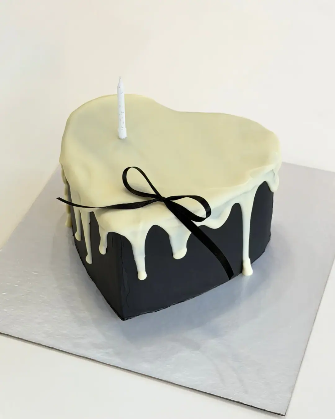 Black heart-shaped cake with a white chocolate drip, a black ribbon bow and a single white candle, a custom cake by Cakes By Vjollca in Pristina, Kosovo