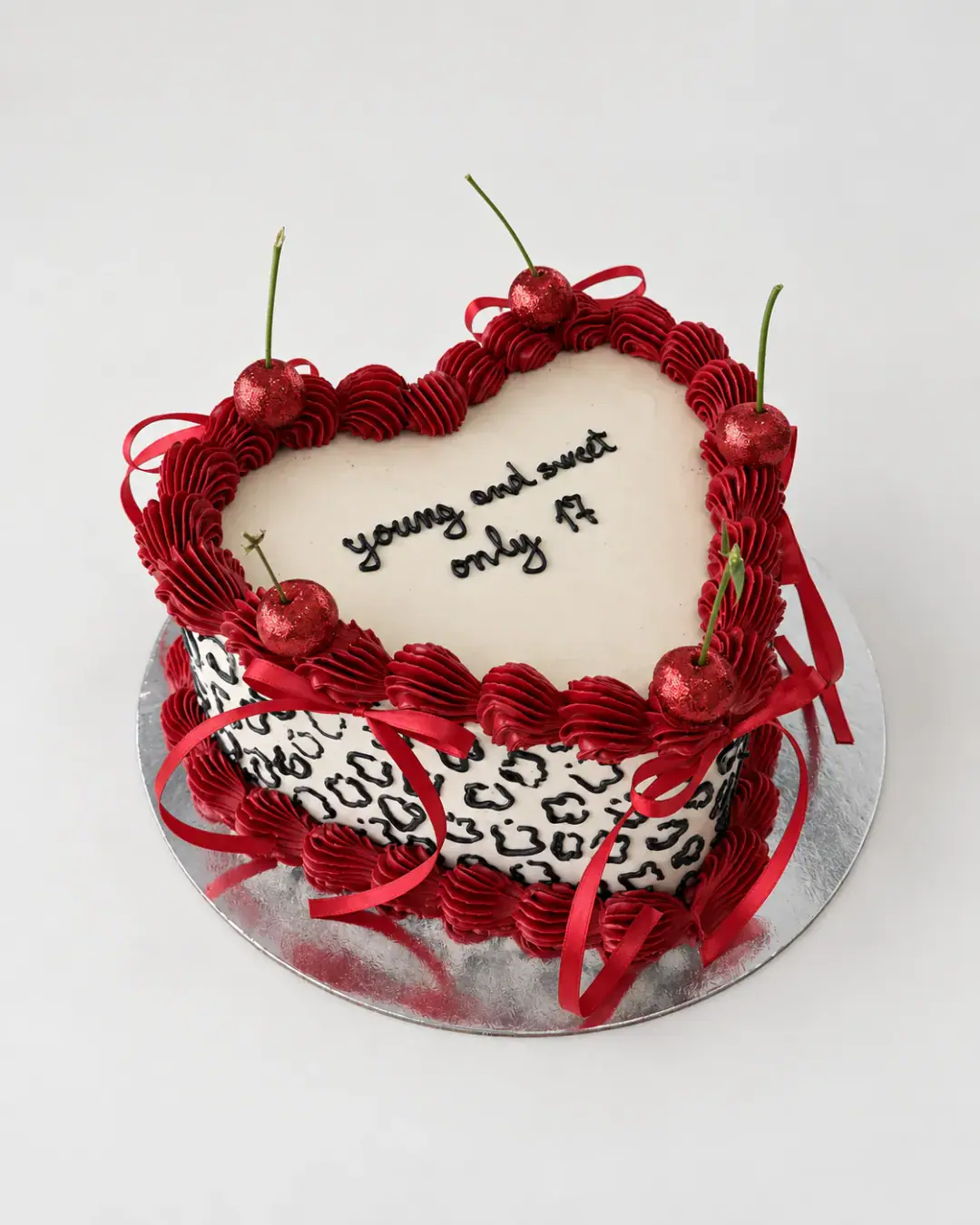 Vintage heart cake with leopard print sides, red piped borders, glittered cherries, red ribbons and 'young and sweet only 17' written on top, a custom cake by Cakes By Vjollca in Pristina, Kosovo