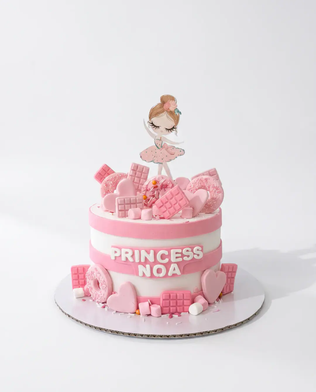 Pink and white princess birthday cake with a ballerina topper and fondant chocolate bars, doughnuts, hearts and marshmallows, a custom cake by Cakes By Vjollca in Pristina, Kosovo