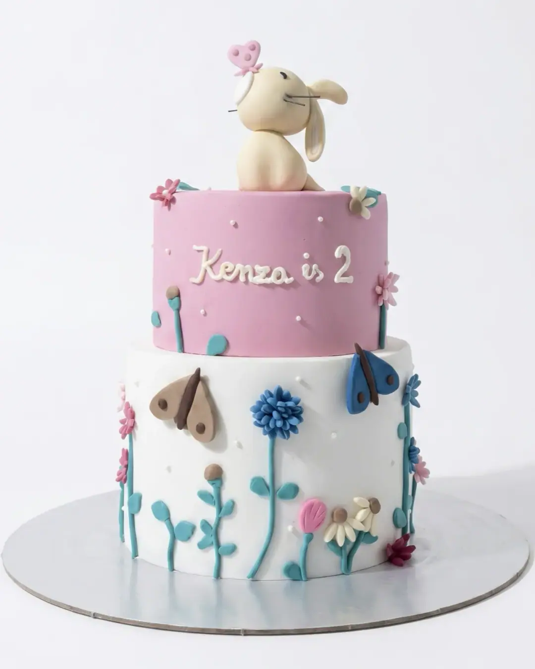 Two-tier pink second birthday cake with a bunny topper and fondant wildflowers, a custom cake by Cakes By Vjollca in Pristina, Kosovo