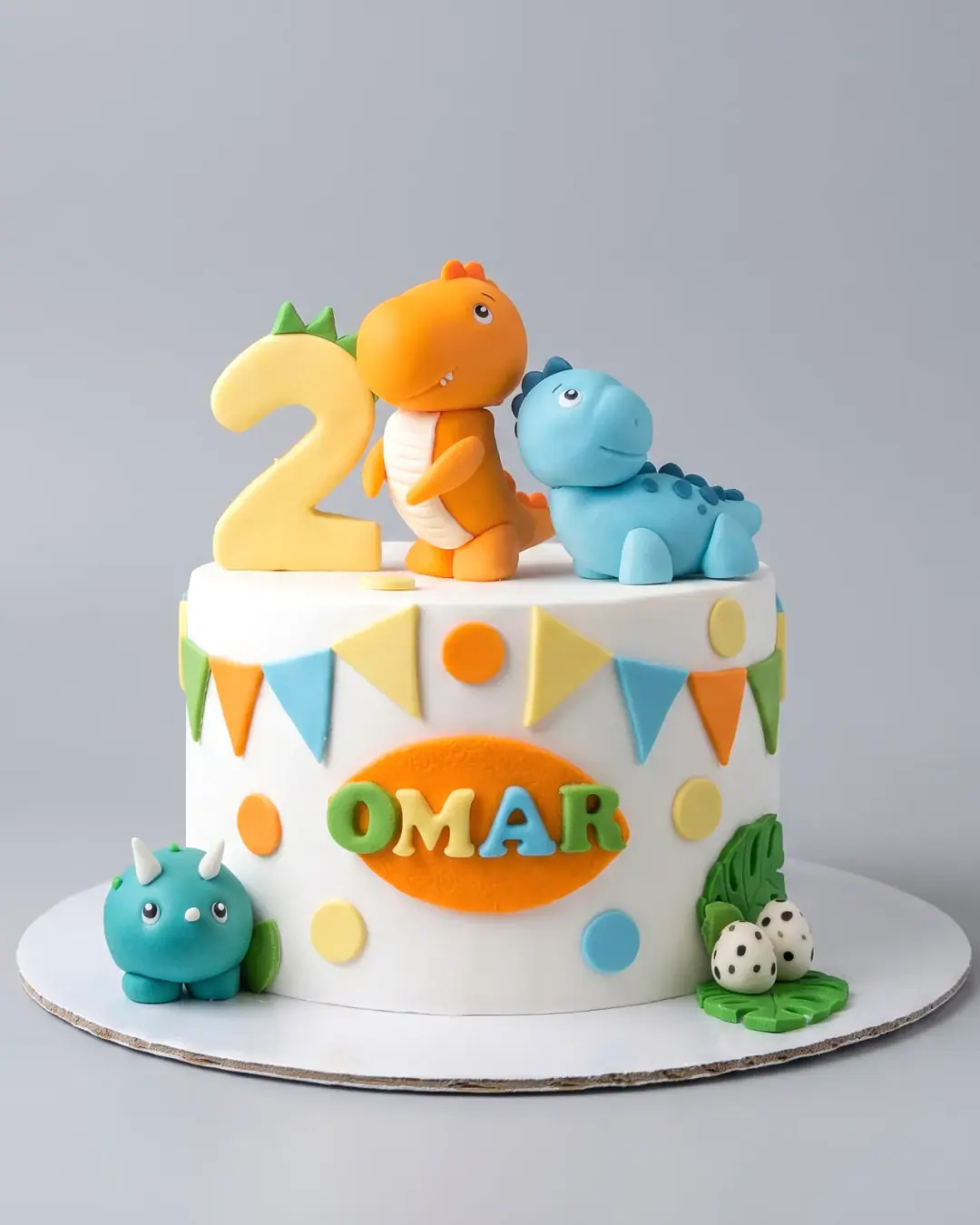 White dinosaur birthday cake with three fondant dinosaurs, a number two, bunting, spotted eggs and the child's name, a custom cake by Cakes By Vjollca in Pristina, Kosovo