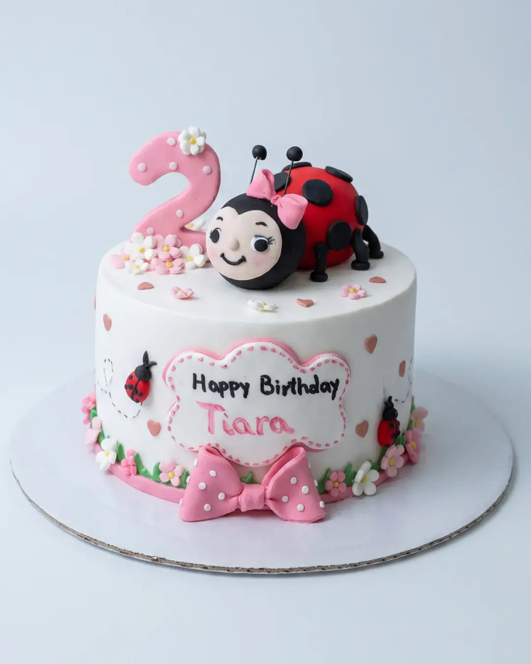 White ladybug birthday cake with a fondant ladybug in a bow, a pink number two, small flowers, hearts and a polka-dot bow, a custom cake by Cakes By Vjollca in Pristina, Kosovo