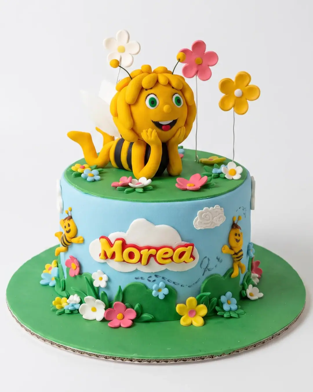 Maya the Bee birthday cake with a fondant Maya on a green meadow top, sky-blue sides, clouds, bees and flowers, a custom cake by Cakes By Vjollca in Pristina, Kosovo