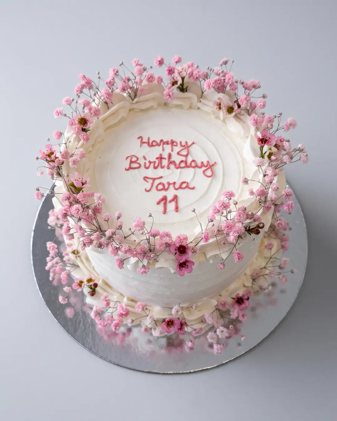 White birthday cake ringed with pink baby's breath and wax flowers, with a piped pink birthday message and the child's name and age, a custom cake by Cakes By Vjollca in Pristina, Kosovo