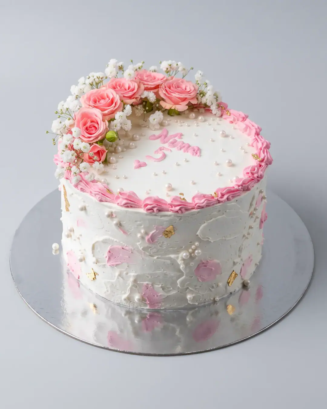 White fifth birthday cake with a crescent of fresh pink roses and baby's breath, pink brushstrokes, gold leaf, pearls and the child's name piped in pink, a custom cake by Cakes By Vjollca in Pristina, Kosovo