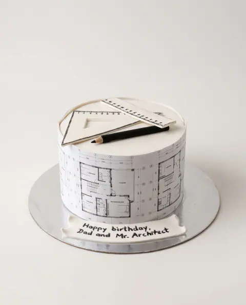 White birthday cake wrapped in printed architectural floor plans, with a fondant set square, ruler and pencil on top and a 'Happy birthday, Dad and Mr. Architect' plaque, a custom cake by Cakes By Vjollca in Pristina, Kosovo