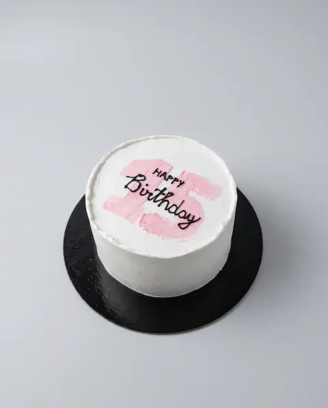 Small white birthday cake with a brushed pink number 15 on top and 'Happy Birthday' piped over it in black script, a custom cake by Cakes By Vjollca in Pristina, Kosovo