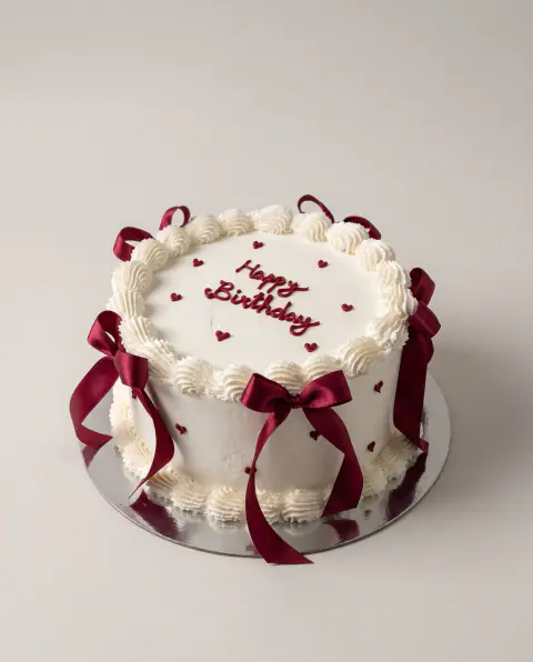 White vintage birthday cake with piped cream borders, burgundy satin bows around the sides and 'Happy Birthday' with small hearts in burgundy script, a custom cake by Cakes By Vjollca in Pristina, Kosovo