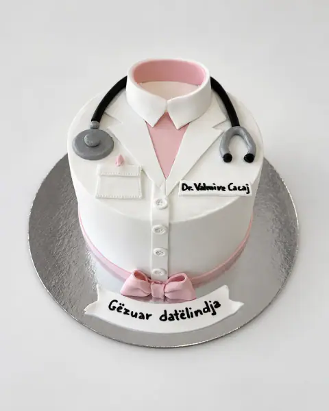 White birthday cake dressed as a doctor's coat with a pink shirt collar, fondant stethoscope, name badge, buttons and pink bow, and a 'Gëzuar ditëlindja' banner, a custom cake by Cakes By Vjollca in Pristina, Kosovo