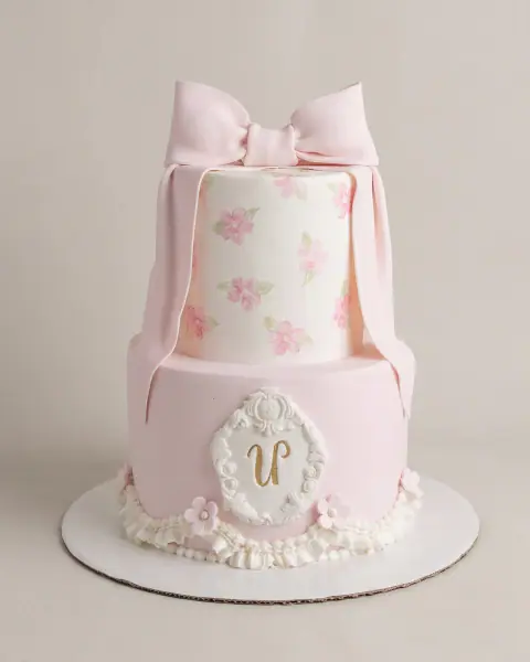 Two-tier pale pink birthday cake with a fondant bow, hand-painted flowers and a gold monogram in a baroque frame, a custom cake by Cakes By Vjollca in Pristina, Kosovo
