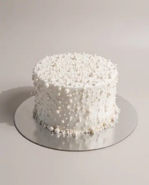 Torte e lartë e bardhë e mbuluar me perla sheqeri të madhësive të ndryshme, me perla më të mëdha të mbledhura në bazë, torte me porosi nga Cakes By Vjollca në Prishtinë