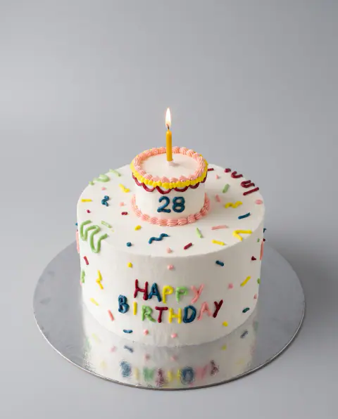 Torte ditëlindjeje me krem të bardhë me spërkatje shumëngjyrëshe të pipuara dhe mbishkrimin 'Happy Birthday', me një torte të vogël me numrin 28 dhe qiri të ndezur sipër, torte me porosi nga Cakes By Vjollca në Prishtinë