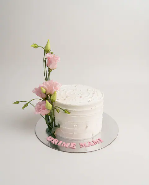 White textured buttercream cake with a tall spray of fresh pink lisianthus at one side and 'Urime Mami' in pink letters on the board, a custom cake by Cakes By Vjollca in Pristina, Kosovo