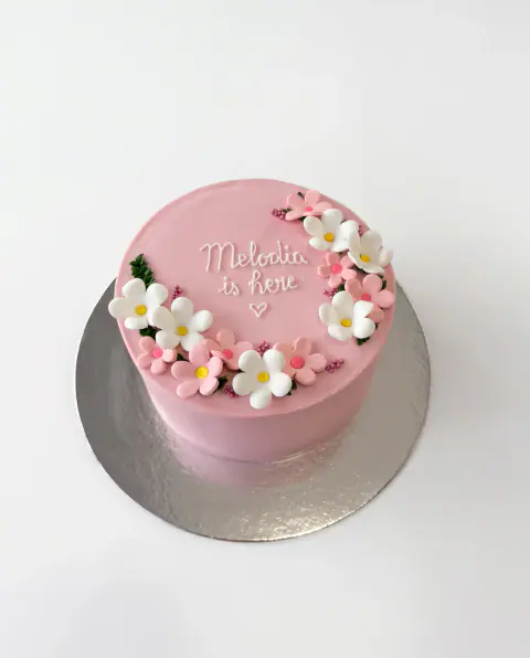 Torte rozë për mirëseardhjen e një bebeje, me emrin e bebes dhe fjalët 'is here' me shkrim të bardhë dhe një gjysmëhënë lulesh sheqeri të bardha e rozë, torte me porosi nga Cakes By Vjollca në Prishtinë