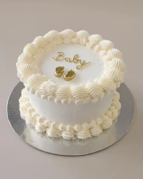 White vintage-style celebration cake with 'Baby' in gold script and two gold baby feet, a custom cake by Cakes By Vjollca in Pristina, Kosovo