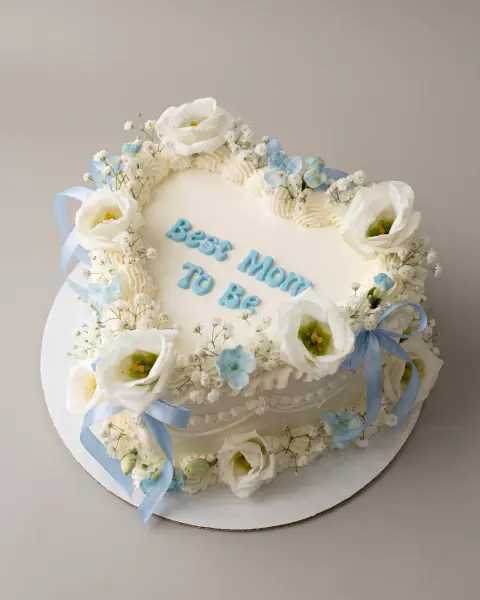 White heart-shaped celebration cake lettered 'Best Mom To Be' with fresh white lisianthus, baby's breath and pale blue ribbons, a custom cake by Cakes By Vjollca in Pristina, Kosovo