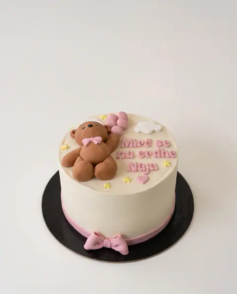 Cream cake welcoming a new baby, with a fondant teddy bear holding pink balloons, 'Mirë se na erdhe' and the baby's name in pink, and a pink ribbon and bow at the base, a custom cake by Cakes By Vjollca in Pristina, Kosovo