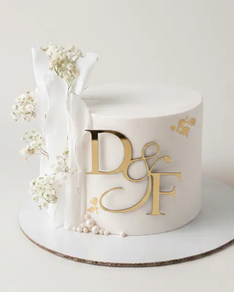 Single-tier white engagement cake with a gold initials topper, draped wafer paper, baby's breath, gold leaf and sugar pearls, a custom cake by Cakes By Vjollca in Pristina, Kosovo