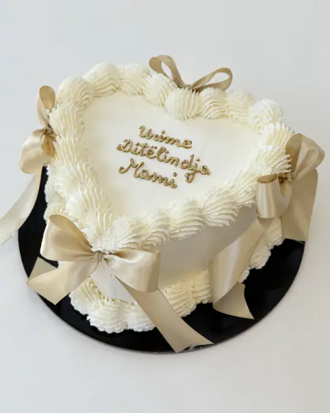 Cream heart-shaped cake with champagne satin bows and piped lettering, a custom cake by Cakes By Vjollca in Pristina, Kosovo