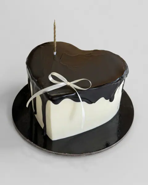White heart-shaped cake with a glossy dark chocolate drip, a white ribbon bow and a single gold candle, a custom cake by Cakes By Vjollca in Pristina, Kosovo