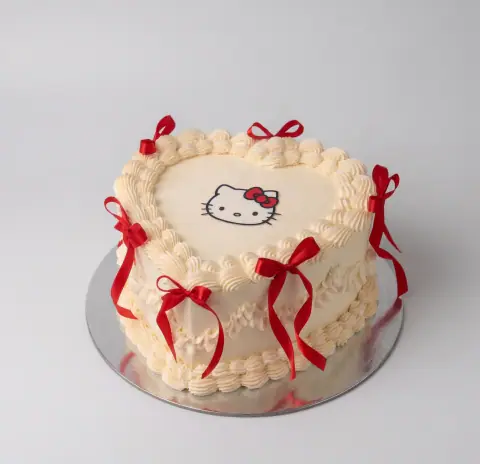 Torte vintage zemër në krem me bordura guackash dhe girlanda të pipuara, fytyrën e Hello Kitty sipër dhe fjongo satine të kuqe rreth anëve, torte me porosi nga Cakes By Vjollca në Prishtinë