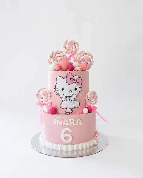 Two-tier pink Hello Kitty birthday cake with pink and white swirl lollipops, candy balls, the child's name and the number 6, a custom cake by Cakes By Vjollca in Pristina, Kosovo