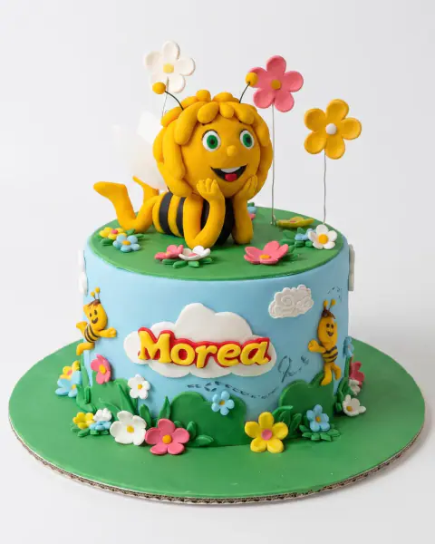 Maya the Bee birthday cake with a fondant Maya on a green meadow top, sky-blue sides, clouds, bees and flowers, a custom cake by Cakes By Vjollca in Pristina, Kosovo