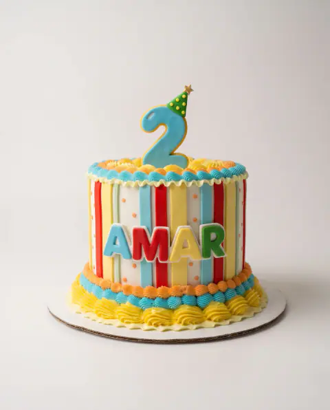 Birthday cake with vertical red, yellow, blue and white stripes, colourful piped borders, the child's name in fondant letters and a blue number 2 in a party hat, a custom cake by Cakes By Vjollca in Pristina, Kosovo