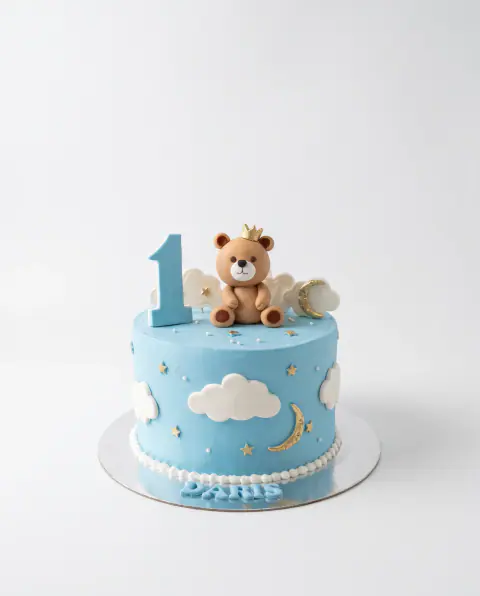Sky-blue first birthday cake with a crowned fondant teddy bear, a blue number 1, fondant clouds, a gold moon and stars and the child's name at the base, a custom cake by Cakes By Vjollca in Pristina, Kosovo