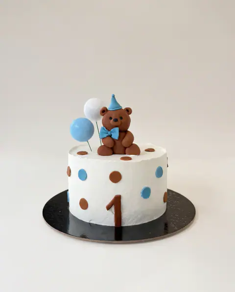 White first birthday cake with brown and blue polka dots, a fondant teddy bear in a party hat and bow tie, blue balloons and a brown number 1, a custom cake by Cakes By Vjollca in Pristina, Kosovo